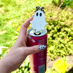 May include: A white ghost-shaped beverage can opener with a leopard print bow attached to the top. The opener is inserted into the tab of a pink Red Bull Peach edition can, featuring visible nutritional information and a barcode on the side.