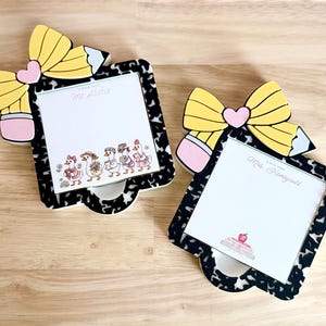 Puede incluir: Dos sujetapapeles decorativos con diseño de lápiz y lazo. Los sujetapapeles tienen un patrón en blanco y negro, y los blocs de notas presentan ilustraciones de gansos y un pastel. Los textos "Ms. Alahia" y "Mrs. Honeycutt" son visibles.