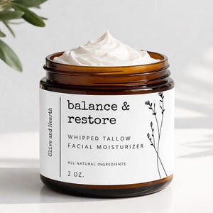 Puede incluir: Un tarro de vidrio marrón lleno de crema hidratante facial de sebo batido blanco. La etiqueta dice "balance & restore" y "Olive and Hearth". El tarro contiene 57 gramos de producto y tiene un diseño minimalista con ilustraciones botánicas.