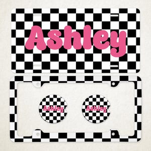 Pode incluir: Uma placa de matrícula retangular e uma moldura correspondente, ambas com um padrão xadrez preto e branco. A placa exibe o nome Ashley em letras cor-de-rosa grandes. Dois porta-copos circulares para carro apresentam o mesmo padrão.