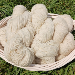 May include: A white wicker basket filled with several skeins of off-white yarn. The yarn is thick and appears to be made of wool. The basket is sitting on a bed of green grass.
