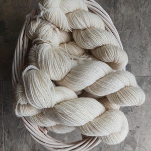 May include: A basket filled with off-white yarn skeins. The yarn is thick and twisted, with a soft, textured appearance. The basket is made of woven, light-colored wicker. The yarn is piled high, suggesting a generous amount for crafting projects.