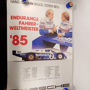 May include: A vintage poster advertising the 1985 World Endurance Championship. The poster features a blue and white Porsche 956 race car with the number 2 on the side. The car is driven by Hans-Joachim Stuck and Derek Bell. The poster also includes the names of the other drivers and their cars in the championship.