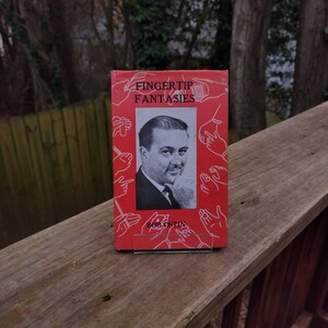 May include: A red book titled "Fingertip Fantasies" by Bob Ostin. The book cover features a black and white photo of a man with a serious expression. The cover is decorated with illustrations of hands performing magic tricks.