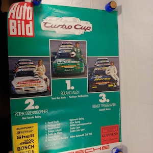 May include: A green and white poster advertising the Turbo Cup race. The poster features three Porsche race cars with the drivers' names and sponsors listed. The text on the poster includes the names of the sponsors, such as Shell, Bosch, and Blaupunkt.