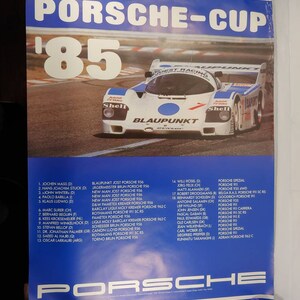 Może przedstawiać: Niebiesko-biały plakat reklamujący serię wyścigów Porsche Cup z 1985 roku. Na plakacie widnieje biały samochód wyścigowy Porsche z niebieskim paskiem i napisami "Blaupunkt" i "Shell" na boku. Plakat zawiera również listę nazwisk kierowców i ich samochodów.