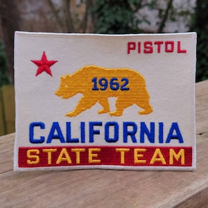 May include: A white patch with red, yellow, and blue embroidery. It features a red star, a yellow bear with the number "1962" on its chest, and the text "PISTOL CALIFORNIA STATE TEAM".