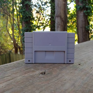 May include: A gray Super Nintendo Entertainment System (SNES) game cartridge. The cartridge is sitting on a wooden surface.