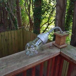 May include: A weathered wooden deck railing with a metal outdoor light fixture attached. The light fixture has a clear glass bulb and a metal arm extending out. The light is not turned on.