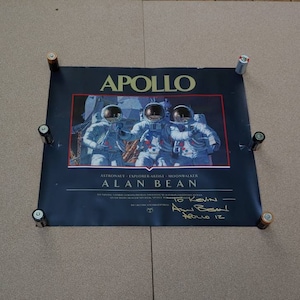 Puede incluir: Un póster negro y azul con una imagen de tres astronautas con trajes espaciales. El póster tiene el título "APOLLO" y el texto "ASTRONAUT - EXPLORER - ARTIST - MOONWALKER ALAN BEAN". El póster está firmado "To Kevin - Alan Bean APOLLO 12".