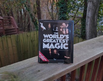 Może przedstawiać: Czarna książka zatytułowana "The World's Greatest Magic" z obrazkiem maga trzymającego talię kart. Okładka książki zawiera również tekst "32 Master Magicians Performing Their Most Mystifying Feats" i "Great Magic Secrets Revealed".