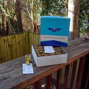 May include: A box of Eagle brand specification grade wiring devices. The box is open and contains several gold-colored electrical components. The box is sitting on a wooden surface.