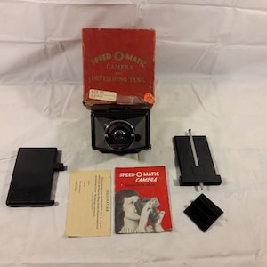 Może przedstawiać: Czarny aparat Speed-O-Matic z czerwonym pudełkiem, instrukcją i zbiornikiem do wywoływania. Pudełko zawiera tekst "Speed-O-Matic Camera Developing Tank" i "$2.98".