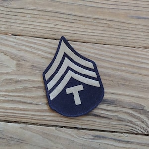 Puede incluir: Un parche de tela azul oscuro con tres rayas blancas en forma de chevron y una letra "T" blanca en el centro. El parche es una insignia de rango militar.