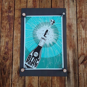 May include: A black framed poster with a turquoise background featuring a bottle of champagne with a woman popping out of the top. The text "Bloc Party" is printed on the bottle. The poster is mounted on a wooden surface.