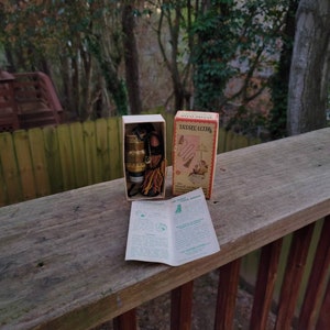 May include: An open box with a brass-colored filter and accessories, a small box with the text "TASSEL FILTER", and an open pamphlet. The items are on a wooden surface with a blurred background of trees and a fence.