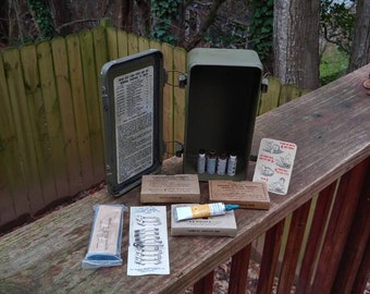 May include: A green metal first aid kit with a hinged lid open, revealing a list of contents and several small tubes. The kit is on a wooden deck with various first aid supplies, including safety pins, gauze, and a tube of antiseptic.