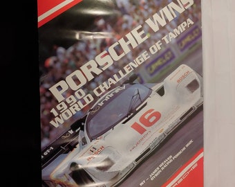 May include: A white Porsche 962C race car with the number 16 on the side. The car is on a track with a crowd of people in the background. The poster reads "Porsche Wins 1990 World Challenge of Tampa".