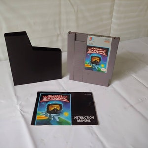 May include: A gray Nintendo Entertainment System (NES) game cartridge with a black label that reads "Captain Skyhawk". The cartridge is in a black plastic case. The game's instruction manual is also included.