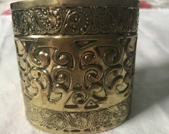 以下が含まれることがあります： 複雑なエンボス加工が施された金色の金属製の容器。容器は丸い形をしていて、口が広い。