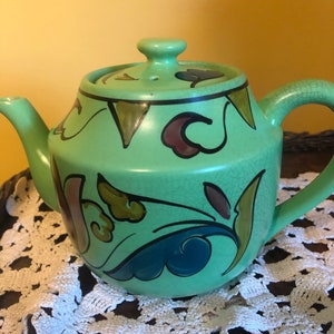 May include: A green ceramic teapot with a floral design in shades of blue, brown, and gold. The teapot has a lid with a small hole in the top.