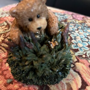 May include: A small brown teddy bear figurine sitting in a bed of green grass with a butterfly on a leaf. The bear is looking to the right and has a brown nose and black eyes.