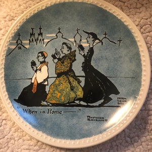 May include: A blue and white ceramic plate with a cartoon illustration of three people walking in a city. The plate has a white border with a beaded design. The text "When in Rome..." is printed on the plate. The artist's signature "Norman Rockwell" is printed on the bottom of the plate.