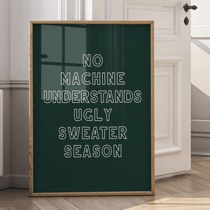 May include: A framed poster with the text "NO MACHINE UNDERSTANDS UGLY SWEATER SEASON" in white letters on a dark green background. The frame is a light brown colour. The poster is leaning against a white door.