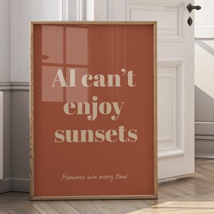 Könnte beinhalten: Ein gerahmter Kunstdruck mit warmem Terrakotta-Hintergrund und hellbeiger Schrift. Der Text lautet "AI can't enjoy sunsets" über "Humans win every time". Der Holzrahmen verleiht dem Kunstwerk eine natürliche Note.