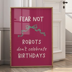 May include: A framed print with a magenta background and a wooden frame. The text "FEAR NOT" is at the top, with a teal bow and the text "ROBOTS don't celebrate BIRTHDAYS" below. The print is displayed near a white door.