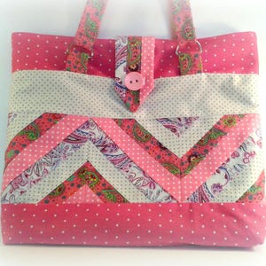 Może przedstawiać: Różowa i biała torba typu tote z patchworku z wzorem chevron. Torba ma różowy guzik i dwa materiałowe paski.