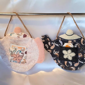 May include: Two fabric tea cozy ornaments in the shape of teapots. One is pink with white paisley print and the other is black with white paisley print. Both have a pocket for tea bags.