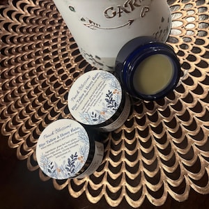 Könnte beinhalten: Eine Sammlung von Peach Blossoms Blue Tallow & Honey Balm-Gläsern und ein blaues Glas mit Balsam. Die Gläser stehen auf einer goldfarbenen, dekorativen, gemusterten Oberfläche. Der Text auf dem Glas lautet "Peach Blossoms Blue Tallow & Honey Balm".