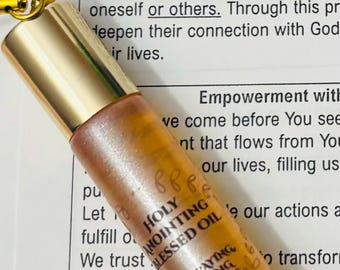 May include: A small, cylindrical bottle of Holy Anointing Blessed Oil with a gold-coloured cap. The bottle has a label with text, set against a backdrop of printed text about prayer and faith.