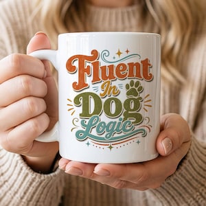 May include: A white ceramic mug featuring the text Fluent In Dog Logic in a retro-style font. The word Fluent is orange, Dog is olive green, and Logic is teal. A small paw print icon and decorative stars and lines surround the text.