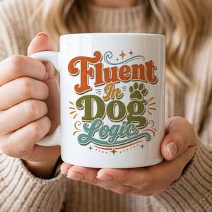 May include: A white ceramic mug featuring the text Fluent In Dog Logic in a retro-style font. The word Fluent is orange, In is yellow, Dog is olive green, and Logic is teal. A small green paw print icon is included next to the text.