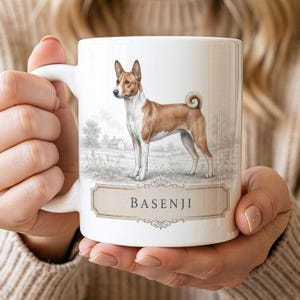 May include: A white ceramic mug featuring a detailed illustration of a brown and white Basenji dog standing in a field. Below the dog, a decorative rectangular label contains the text BASENJI in a dark, classic serif font with scrollwork.