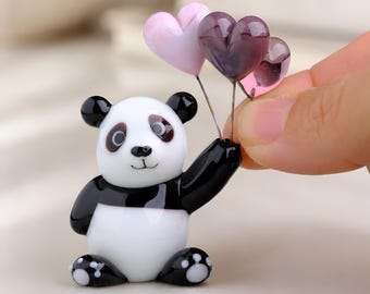 May include: A glass panda figurine in black and white, holding three heart-shaped balloons in shades of pink and purple. The panda has black ears, eyes, and paws. The balloons are attached to thin silver wires.