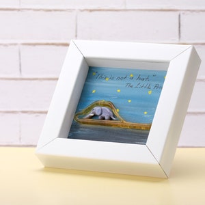 Op de afbeelding: Witte ingelijste afbeelding met een blauwe achtergrond en een citaat uit De Kleine Prins: "Dit is geen hoed." Een olifant is afgebeeld in de afbeelding, rijdend op een klein bootje.