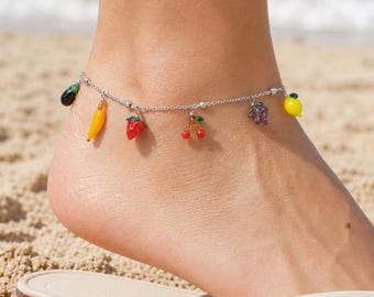 May include: A silver anklet featuring charms shaped like various fruits, such as a banana, strawberry, and cherries. The anklet is displayed on a foot wearing a white flip-flop, with a sandy beach and sea in the background.