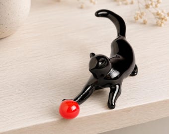 May include: A glossy black cat ornament playing with a small red ball. The cat is in a playful pose, with its front paw extended towards the ball. The ornament is set against a light-coloured wooden surface.