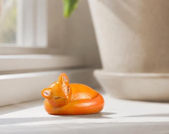 May include: A small, smooth, orange glass figurine of a fox curled into a sleeping position. The fox has closed eyes, pointed ears with white inner details, and a sleek, glossy finish that reflects light, capturing a peaceful, resting posture.
