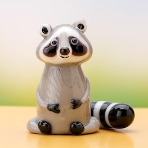 Puede incluir: Figura de cristal de un mapache con cuerpo gris, cara blanca y marcas negras. El mapache está sentado con la cola enrollada detrás. La cola es de rayas blancas y negras. Adorno decorativo.