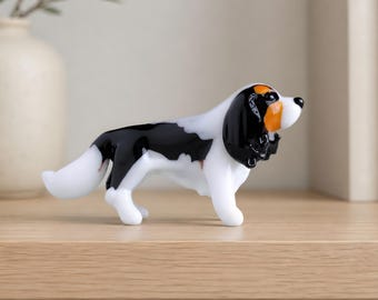 May include: A glass figurine of a Cavalier King Charles Spaniel dog. The dog has a white body with black and orange markings on its head and back. The figurine is standing on a wooden surface.