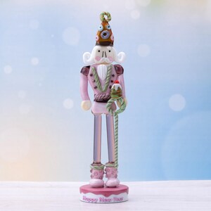 Könnte beinhalten: Dekorative rosa-braune Nussknacker-Figur mit weißem Schnurrbart und hohem Hut mit farbenfrohem Design. Die Figur hält ein grün-weißes Seil und steht auf einem Sockel mit der Aufschrift "Happy New Year".