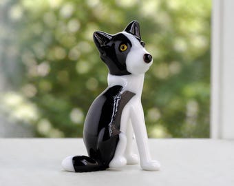 May include: A glass dog figurine in black and white with yellow eyes. The dog is depicted in a seated pose, with a curled tail. The figurine has a glossy finish and appears handcrafted. The background is blurred, suggesting an outdoor setting. The figurine is likely a decorative item.