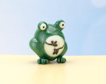 May include: A green glass frog figurine with white and black details. The frog has large, round eyes and a wide, toothy smile.