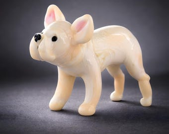 May include: A cream-coloured figurine of a French Bulldog. The dog has black eyes and a black nose. The inside of the ears are pink. The figurine is standing on a dark grey surface.