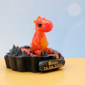 May include: Orange and yellow dragon figurine sitting on a black lava rock base with the name Brian and the date 23.06.2024.