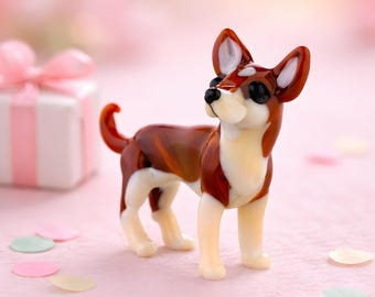 May include: A glass Chihuahua dog figurine, with brown and white detailing. The dog stands on a pink surface, with confetti and a gift box with a pink bow in the background. The dog has black eyes and a curled tail.